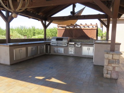 Picture of a stamped concrete patio and concrete countertop installed underneath a patio cover held up by stacked stone pillars. There is a barbecue and sink installed into the countertop